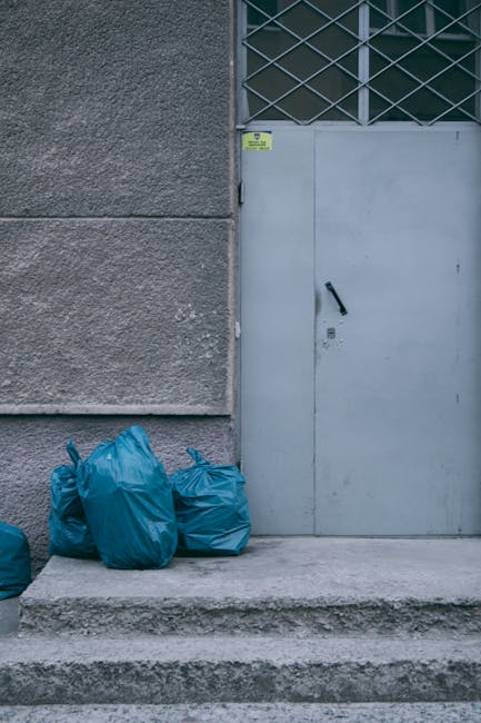 A cluttered scene outside a building shows a large pile of black rubbish bags, some torn or partially open, resting against a textured, weathered beige wall. Among the debris, there are flattened card