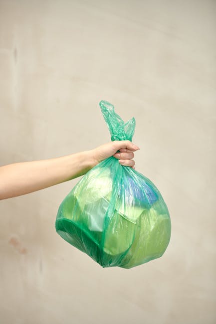 A person's hand and forearm extending from the left side of the image, holding a green plastic rubbish bag with a slightly shiny, crinkled texture. The bag appears to be filled with lightweight waste 