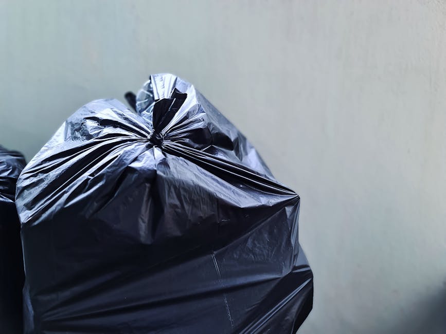 A person's hand and forearm extending from the left side of the image, holding a green plastic rubbish bag with a slightly shiny, crinkled texture. The bag appears to be filled with lightweight waste 
